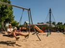 Un papa tenant tendrement son bébé dans ses bras à l'aire de jeux du camping, tous deux pieds nus dans le sable, profitant d'un moment de complicité et de détente en plein air