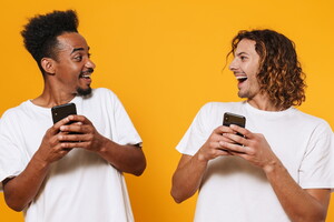 Camping avec application mobile dans les Landes (4) Portrait de deux jeunes hommes heureux tenant leurs téléphones et se regardant isolés dans un fond jaune